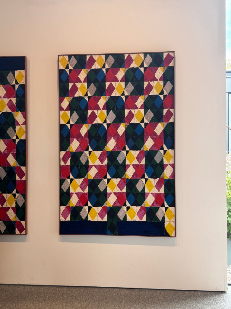 壁に掛けられたカラフルな幾何学模様の絵画。赤、黄、緑、青、白などの色が使用され、ダイヤモンド形のパターンが繰り返されている。