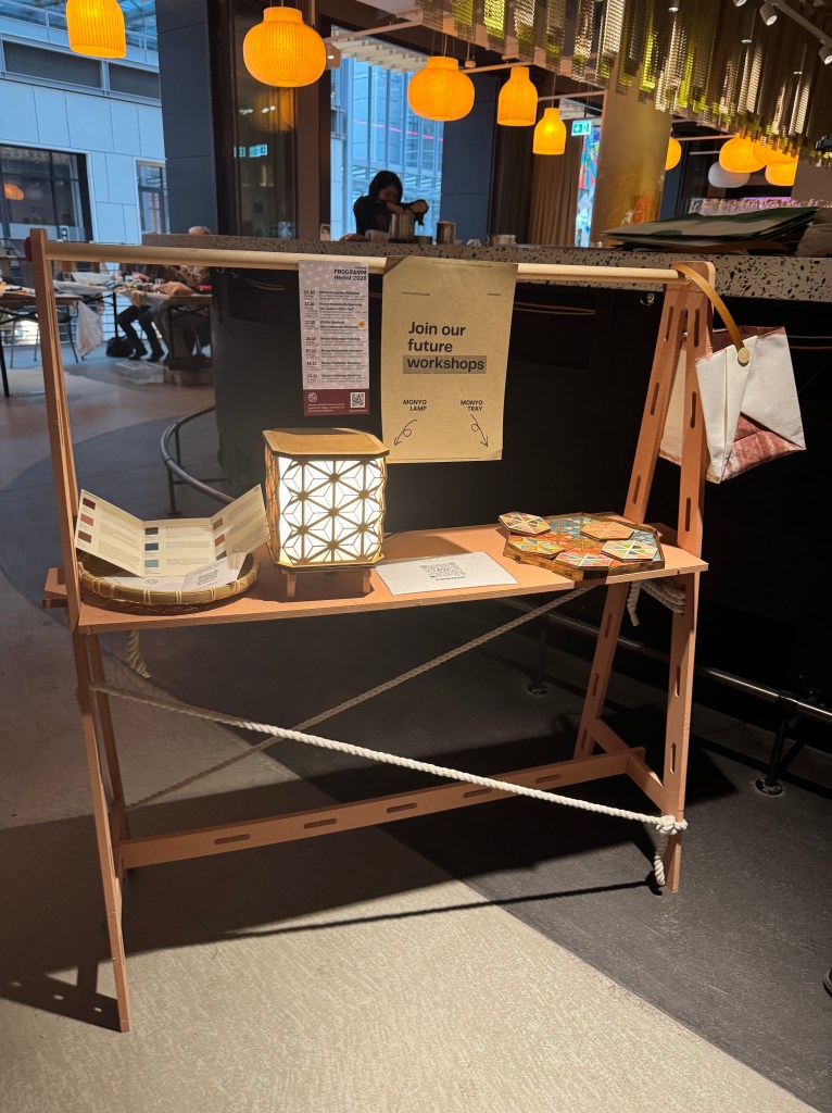 ワークショップの参加を促す案内のあるディスプレイ棚。照明、ランプ、カラーパレット、QRコードが展示されている。